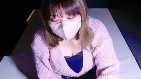 Media: Video of a woman with straight, shoulder-length light brown hair, wearing a pink knitted cardigan, blue lace bra, and a white face mask, sitting indoors.