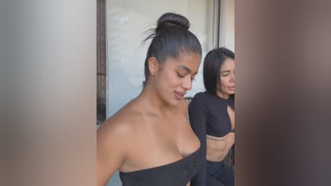 Media: Video of two young women with tan skin and long black hair, wearing black strapless tops, standing side by side in a doorway.