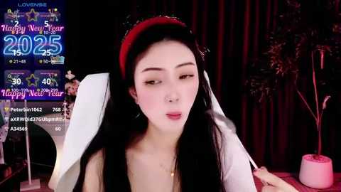 Media: Video of an Asian woman with fair skin, wearing a red hat and white headpiece, sitting at a desk with a \"Happy New Year 2025\" background.