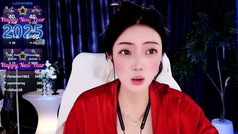 Media: Video of a young Asian woman with fair skin, dark hair in a bun, wearing a red satin robe, sitting in a white chair. Background includes festive decorations, a potted plant, and a TV screen displaying \"Happy New Year 2025.\