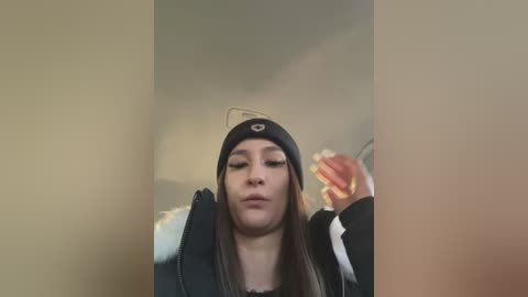 Media: A video of a young woman with long brown hair, wearing a black beanie and black jacket, blowing a kiss with her right hand. Background shows a beige wall and a blurred figure in the corner.