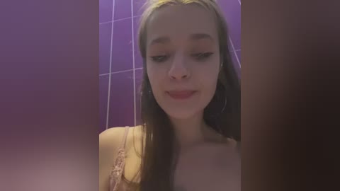 Media: Video of a young Caucasian woman with long, straight brown hair and light skin, wearing a yellow lace top, in a purple-tiled bathroom, smiling softly.