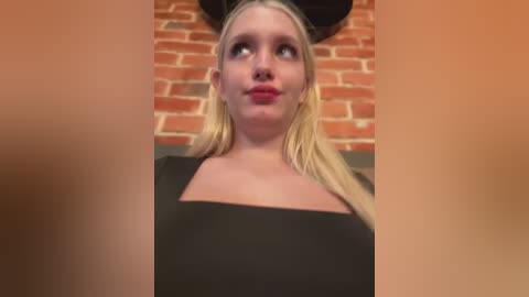 Media: Video of a blonde woman with fair skin, wearing a black dress, against a red brick wall, with a blurred face in the foreground.