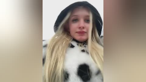 Media: A video of a young woman with long, blonde hair in a black cow-patterned hoodie, wearing a neutral expression, with blurred, soft-focus background.