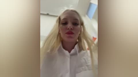 Media: A video of a blonde woman with fair skin, wearing a white button-up shirt, stands indoors with a blurred background, looking forward.