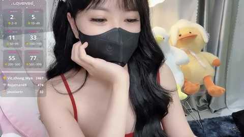 Media: Video of an East Asian woman with long black hair, wearing a black face mask, red bra, and a white top, sitting near stuffed ducks in a cozy room.