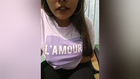 Media: A video of a young woman with medium brown skin and long dark hair, wearing a white t-shirt with \"LAMOUR\" in purple text and black leggings, sitting in a chair.