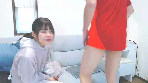 Media: Video of an East Asian woman with dark hair, wearing a lavender hoodie, kneeling on a light blue couch, watching a red-clad woman from behind.