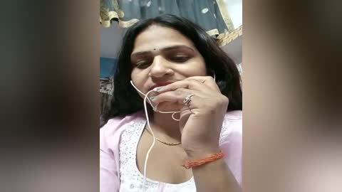 Media: A video of a South Asian woman with long black hair, wearing a pink cardigan, listening to music with earphones, in a home setting.