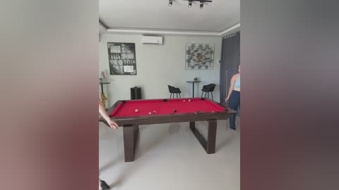 Media: A video of a modern, minimalist room with a red pool table, black chairs, gray walls, and framed artwork; a woman in a blue tank top stands nearby.