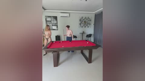 Media: Video of a modern, bright living room with a pool table, two women playing pool, one topless, one in jeans.