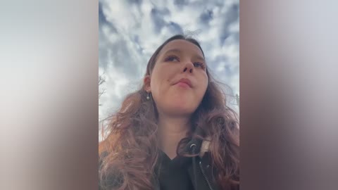 Media: Video of a young woman with wavy, shoulder-length auburn hair, wearing a black jacket, looking up at a cloudy sky.