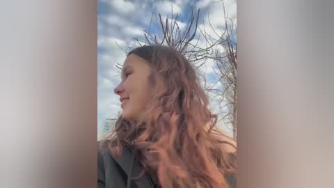 Media: Video of a young woman with long, wavy brown hair, wearing a gray coat, gazing upwards against a cloudy sky.