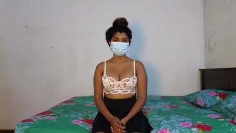 Media: Video of a South Asian woman with medium brown skin, wearing a white lace bra, black skirt, and blue face mask, sitting on a green floral bedspread in a simple room with white walls.