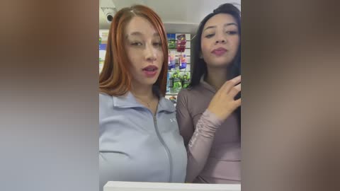 Media: Video of two women in a store, one with light skin and red hair in a light blue zip-up, the other with tan skin and black hair in a pink top. Background shows colorful product displays.