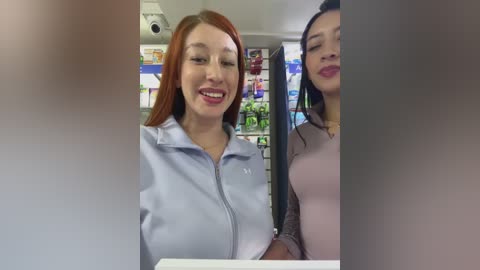 Media: Video of two women smiling in a convenience store. One with red hair in a light blue jacket, the other with long black hair in a pink sweater. Background shows colorful product displays.