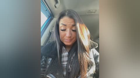 Media: A video of a Latina woman with long, straight, dark hair and a light tan complexion, wearing a black jacket over a plaid shirt, seated in a car with sunlight streaming in.