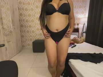 Media: Video of a curvy, light-skinned woman with long black hair in black lingerie, standing in a modern bedroom with patterned wallpaper, a bed, and a lamp.