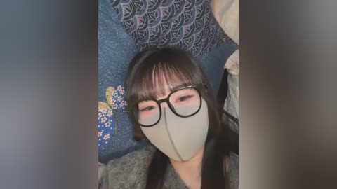 Media: Video of a woman with long black hair, wearing glasses and a grey mask, sitting on a blue patterned cushion with a butterfly embroidery, indoors.