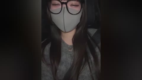 Media: A dimly-lit video of an Asian woman wearing a gray surgical mask, black-framed glasses, and a dark gray hoodie, sitting in a car with dark interior.