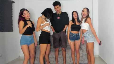 Media: Video of a man in a black t-shirt and shorts, surrounded by four women in casual, revealing outfits. They stand in a tight cluster in a minimalist room.