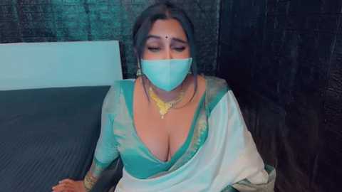 Media: Video of a woman with dark skin, wearing a teal saree, gold jewelry, and a blue mask, sitting on a dark bed, in a dimly lit room with dark walls.