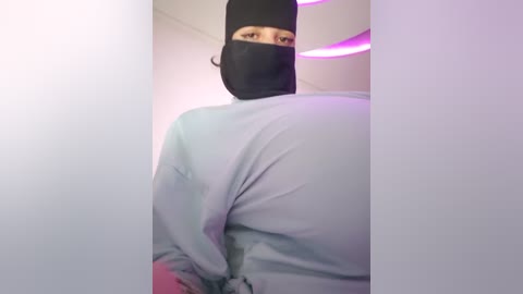 Media: Video of a person with light skin, wearing a black mask and light blue robe, standing in a minimalist room with a purple light.