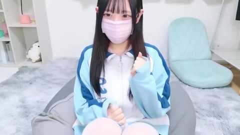 Media: Video of a young East Asian woman in a blue and white tracksuit with a mask, sitting on a gray chair in a pastel-toned room with light blue cushions and a white shelf.