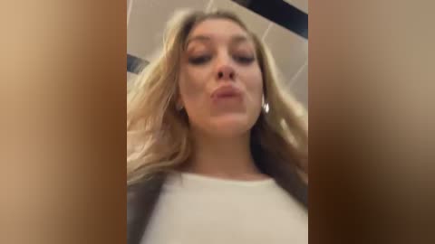 Media: A low-angle video of a young, fair-skinned woman with long blonde hair, wearing a white top and black jacket, puckering her lips in a kiss.