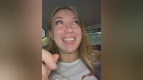 Media: Video of a smiling young woman with straight blonde hair, wearing a white shirt, driving a car with a green sign visible in the background.