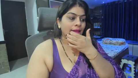 Media: Video of a woman with long black hair, wearing a purple saree and gold jewelry, sitting on a gray chair in a modern bedroom with dark curtains and a bed with white sheets.