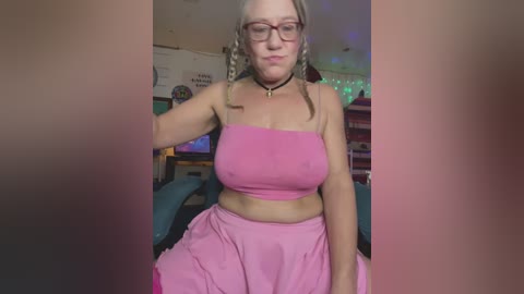 Media: Video of a plus-sized, light-skinned woman with blonde hair in braids, wearing a pink crop top and ruffled skirt, sitting on a blue chair, in a colorful, cluttered room with posters and string lights.