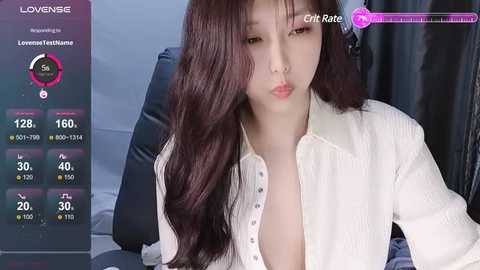 Media: Video of a young Asian woman with long dark hair, wearing a white blouse with a low neckline, sitting on a dark couch in a dimly lit room.