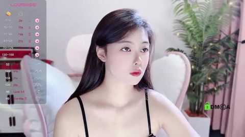 Media: Video of a young Asian woman with long black hair, wearing a black spaghetti-strap top, looking introspective. Background features a white bed, green plant, and pink curtains.