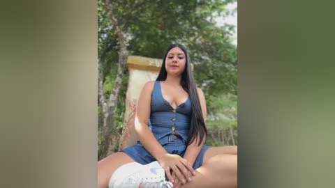 Media: Video of a young Latina woman with long black hair, wearing a blue denim romper, sitting cross-legged outdoors, surrounded by green foliage.