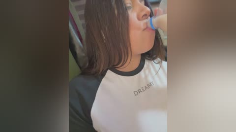 Media: A video captures a young woman with shoulder-length brown hair, wearing a black and white \"Dream\" T-shirt, brushing her teeth with a blue toothbrush. The background is blurred, suggesting an indoor setting.