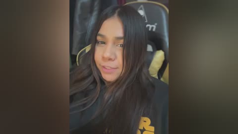 Media: Video of a young woman with long, dark hair, light brown skin, and a warm smile, sitting in a black gaming chair with yellow accents. She wears a black jacket with yellow and white letters.