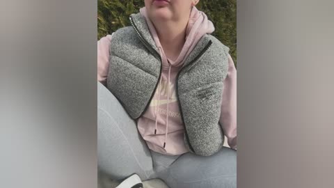 Media: Video of a person wearing a grey quilted vest over a pink hoodie, sitting on a grassy surface, partially blurred background.