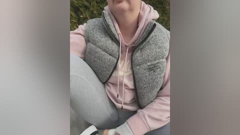 Media: Video of a woman in a gray puffy vest over a pink hoodie, seated outdoors with blurred greenery background.