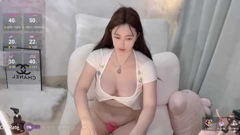 Media: Video of a young East Asian woman with long black hair, wearing a white crop top and no pants, sitting on a white couch in a bedroom.