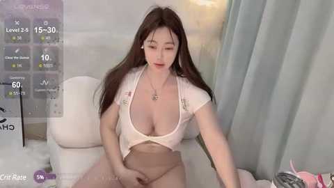 Media: Video of an East Asian woman with long black hair, wearing a low-cut white crop top, revealing ample cleavage, and beige panties, sitting on a white couch. Background includes a soft, light-colored curtain and a digital interface with fitness stats.