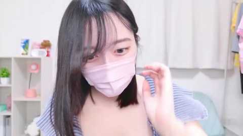 Media: Video of an East Asian woman with straight black hair, wearing a white face mask, adjusting her hair in a room with pastel-colored decor and potted plants.