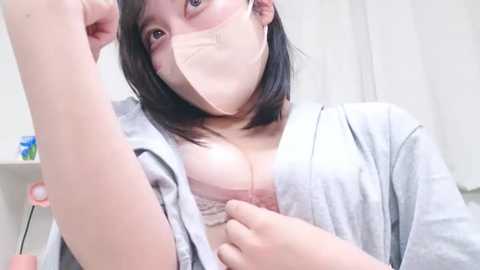 Media: Video of an Asian woman with short black hair and light skin, wearing a face mask, gray cardigan, and revealing a pink bra, exposing cleavage. She's in a brightly lit room with a white curtain background.