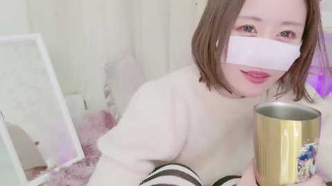 Media: Video of a young Asian woman with short brown hair, wearing a white mask, white sweater, and holding a metallic cup. She sits on a pink carpet in a softly lit, minimalist room.