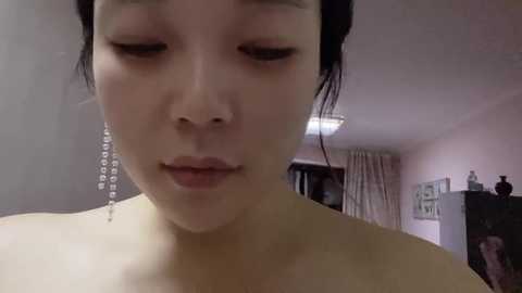 Media: A close-up video of an Asian woman with light skin, dark hair pulled back, wearing long dangling earrings, and looking introspective in a dimly lit room.