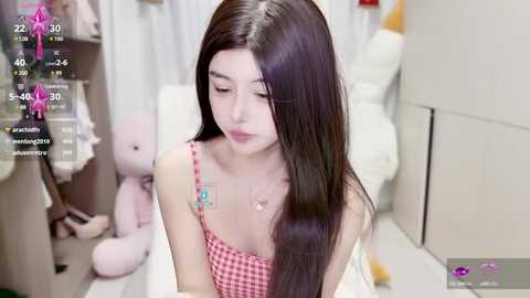 Media: A video of a young Asian woman with long, straight dark hair, wearing a pink and white checkered spaghetti-strap top, sitting in a room filled with plush toys and a white bed.