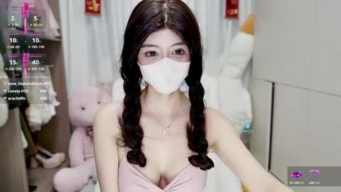 Media: Video of an East Asian woman with long black braided hair, wearing a white face mask and a pink strapless top, standing in a room with stuffed animals and a bed, with a virtual camera overlay.