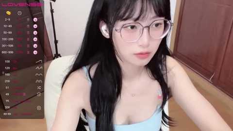 Media: Video of a young Asian woman with long black hair, wearing glasses and a blue tank top, sitting in a dimly lit room.