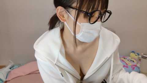 Media: Video of a young Asian woman with glasses, wearing a white face mask and a partially unzipped white hoodie, revealing cleavage. She's indoors, with a stuffed toy and a bed in the background.