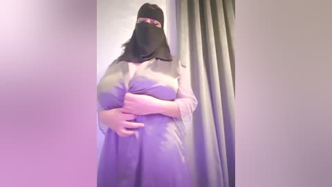 Media: Video of a person wearing a black niqab and a light purple dress, standing against a purple curtain in a dimly lit room.